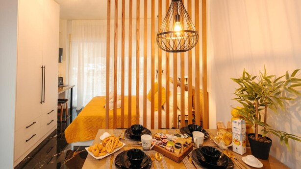 A dining area with a table set for two, a hanging light fixture, a bed with yellow bedding, and a potted plant.