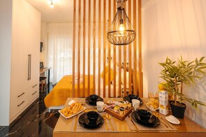 A dining area with a table set for two, a hanging light fixture, a bed with yellow bedding, and a potted plant. - Apartment 'Mantisses 4 Studio' with Balcony, Wi-Fi and Air Conditioning (?a?aµ?ta)