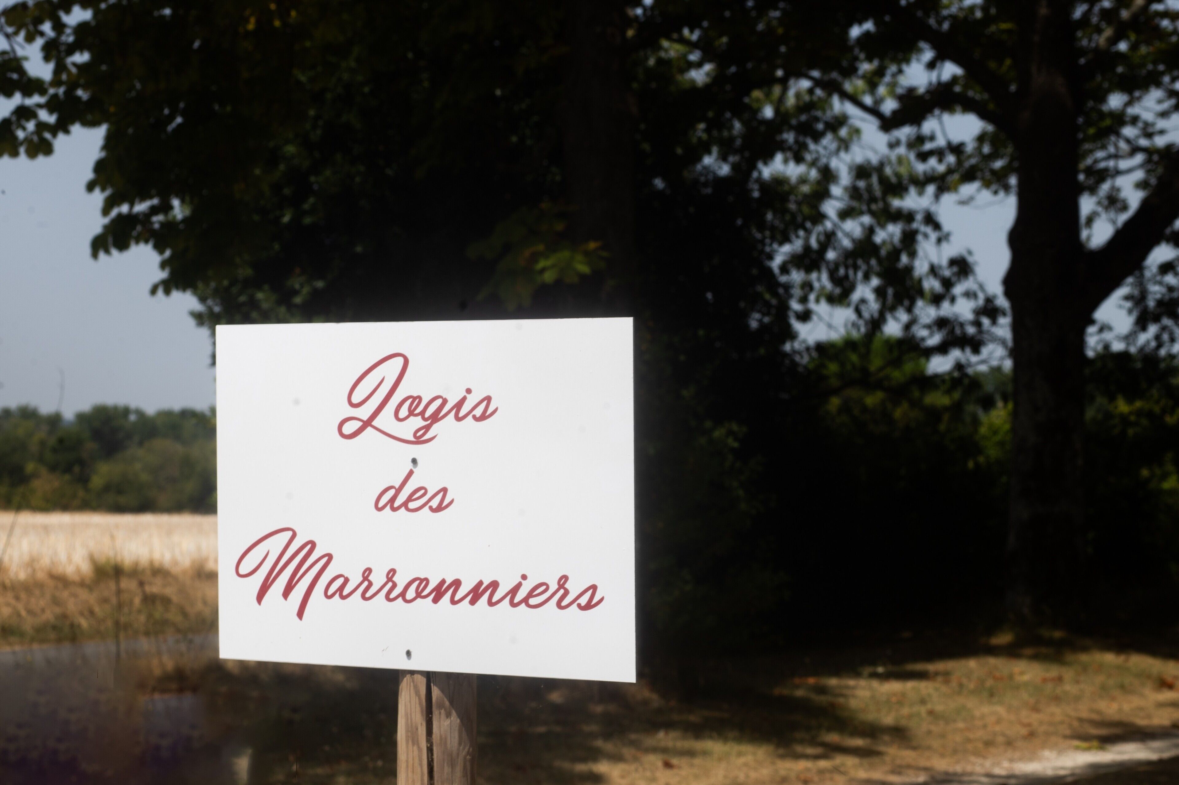 Une enseigne avec l’inscription « Logis des Marronniers » en caractères cursifs, placée dans un cadre naturel avec des arbres et un champ en arrière-plan.