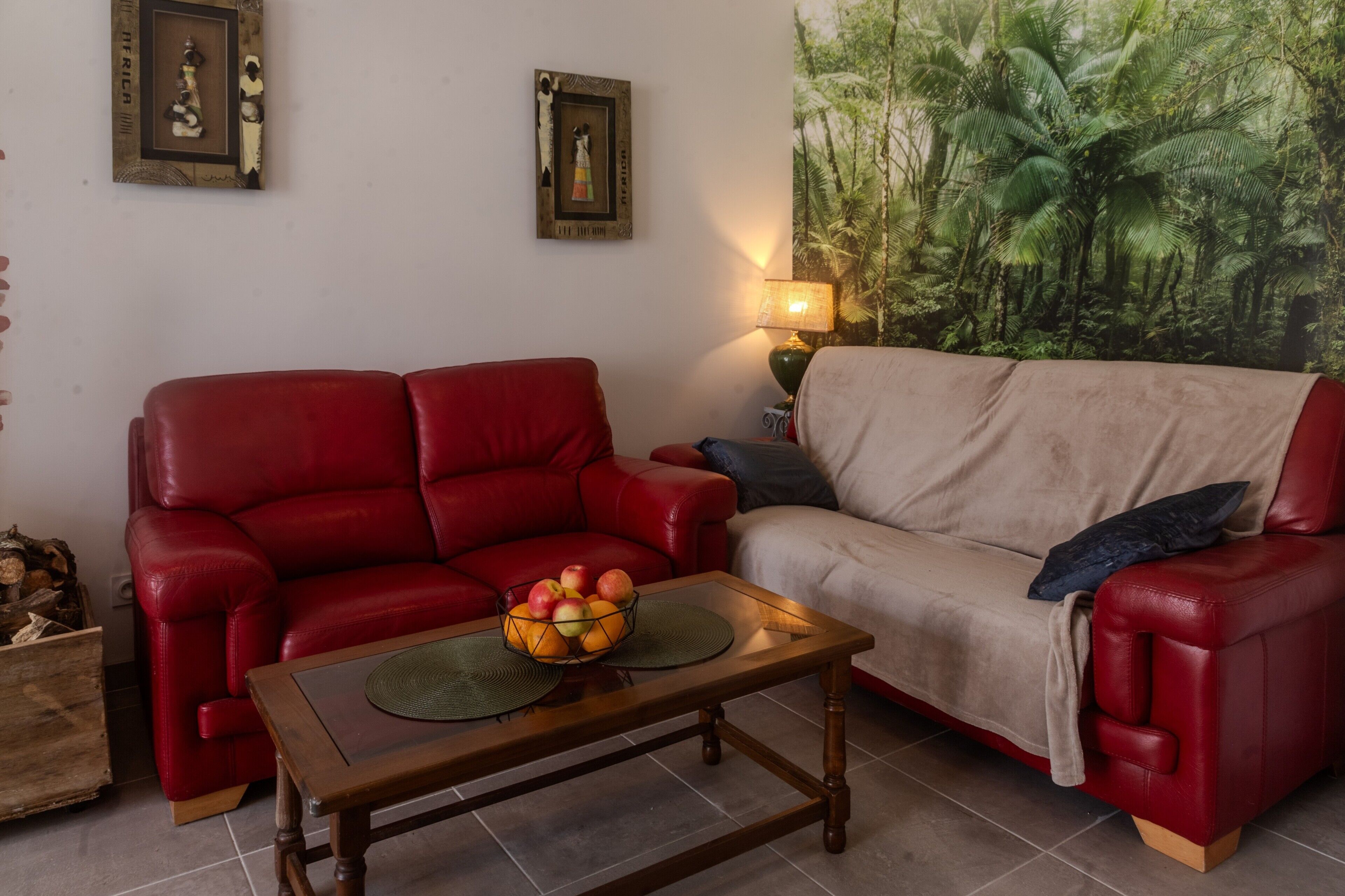A living room with a red leather sofa, a beige sofa, a wooden coffee table with a fruit bowl, and a wall tapestry.