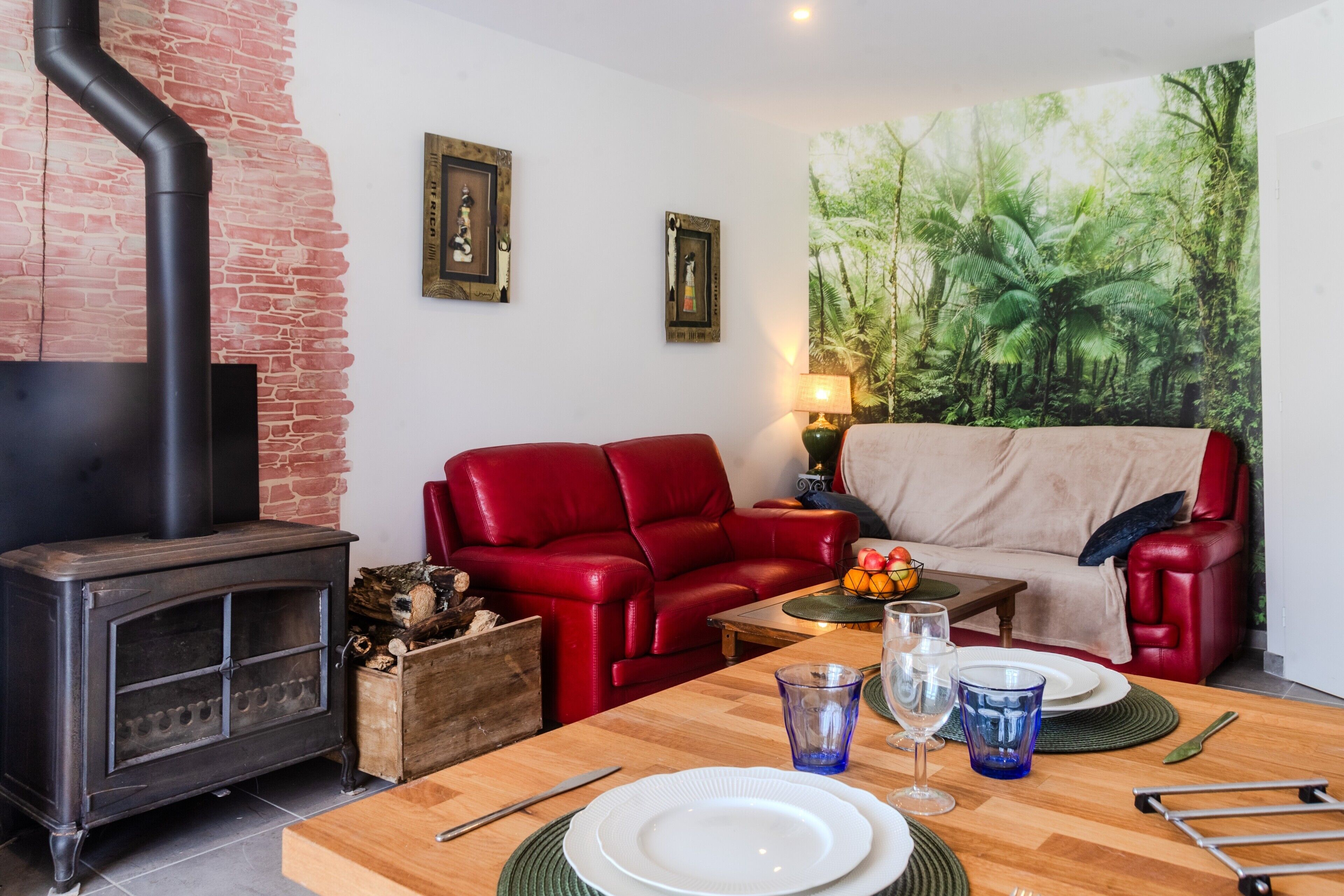 Een woonkamer met een open haard, een rode leren bank, een eettafel met borden en glazen, en een groot muurschildering van een bos.