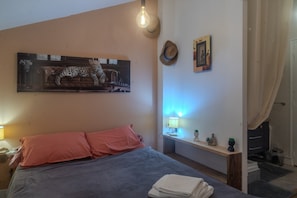 Et soveværelse med seng, puder, et billedværk på væggen, et natbord med lampe og gardin.
