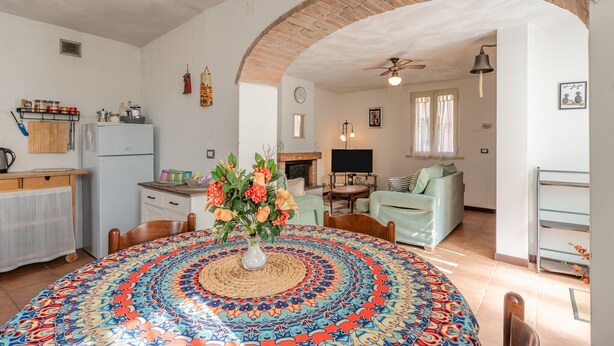 A living room with a fireplace, a dining table with a patterned tablecloth, and a kitchen area with a refrigerator and cabinets.