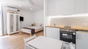 A modern, compact room with a bed, a small kitchenette, a white table, and a TV mounted on the wall. - Apartment 'Portblanc' with Balcony, Wi-Fi, and Air Conditioning (Arenys de Mar)