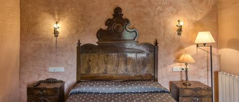 Un dormitorio con cabecera de madera, colcha estampada, dos mesitas de noche con lámparas y un radiador.