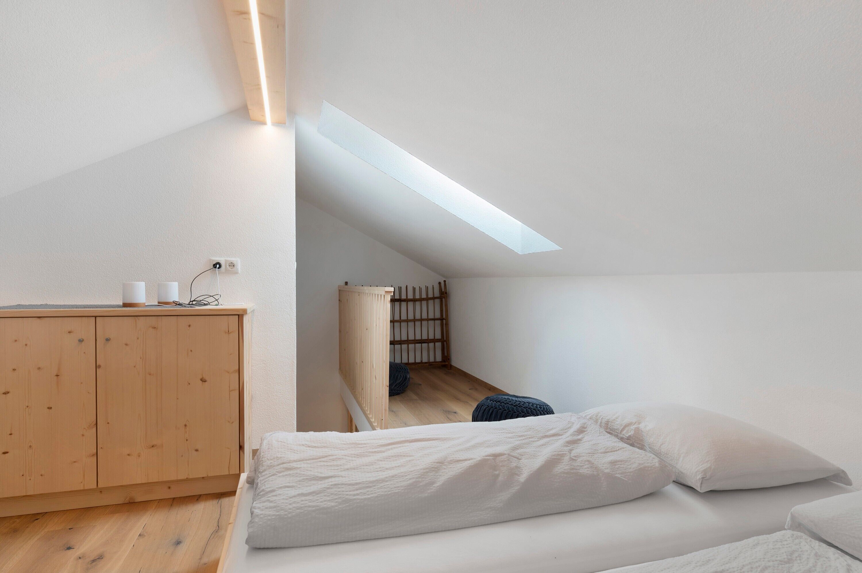Ein schlichter Raum mit schrägen Wänden, einem Holzkabinett, einem Bett mit weißer Bettwäsche und einem Dachfenster.