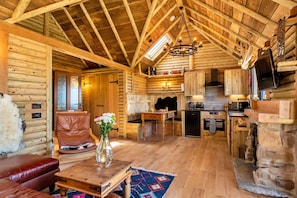 A log cabin interior with a wooden ceiling, a fireplace, a kitchen area, and a seating area with a leather couch and a coffee table.