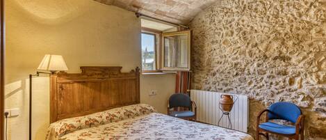Un dormitorio con una cama, dos sillas, un armario de madera, una pared de piedra y una ventana.
