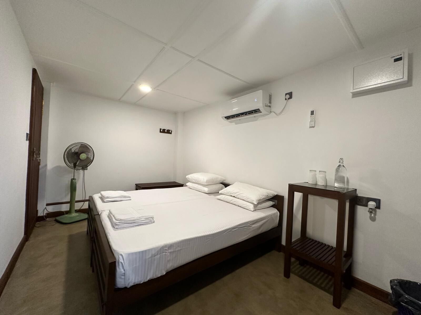 A hotel room with a single bed, a bedside table, a nightstand, a standing fan, an air conditioning unit, and a small table with a bottle and a glass.