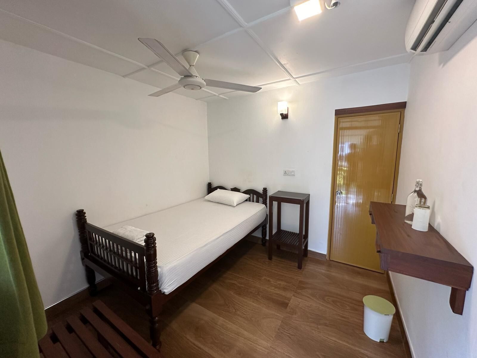 A small, clean room with a single bed, a wooden nightstand, a small table, a wall-mounted shelf, and a door.