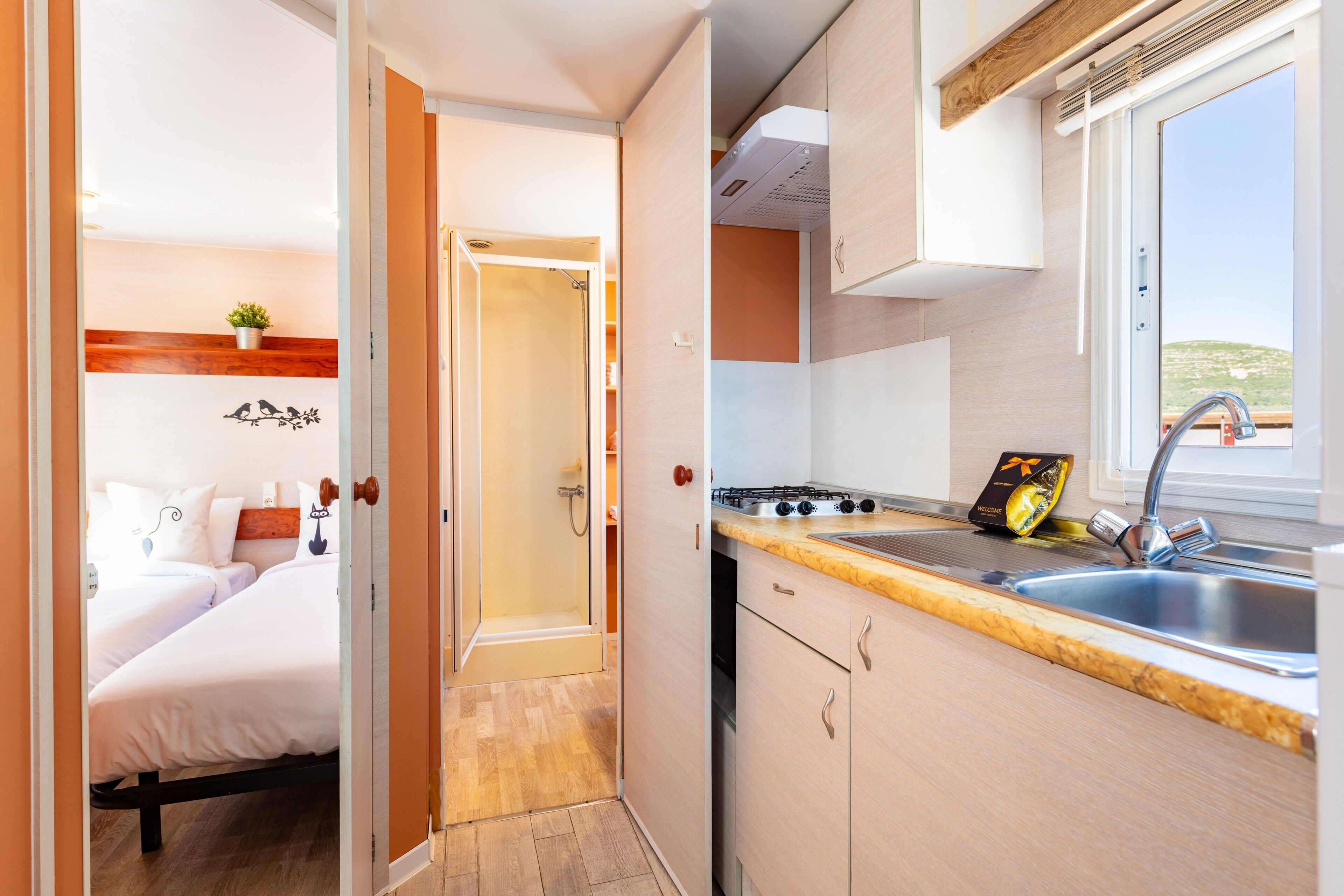 A compact kitchen with a sink, stove, and cabinets. A door leads to a bedroom with a bed and a small plant.