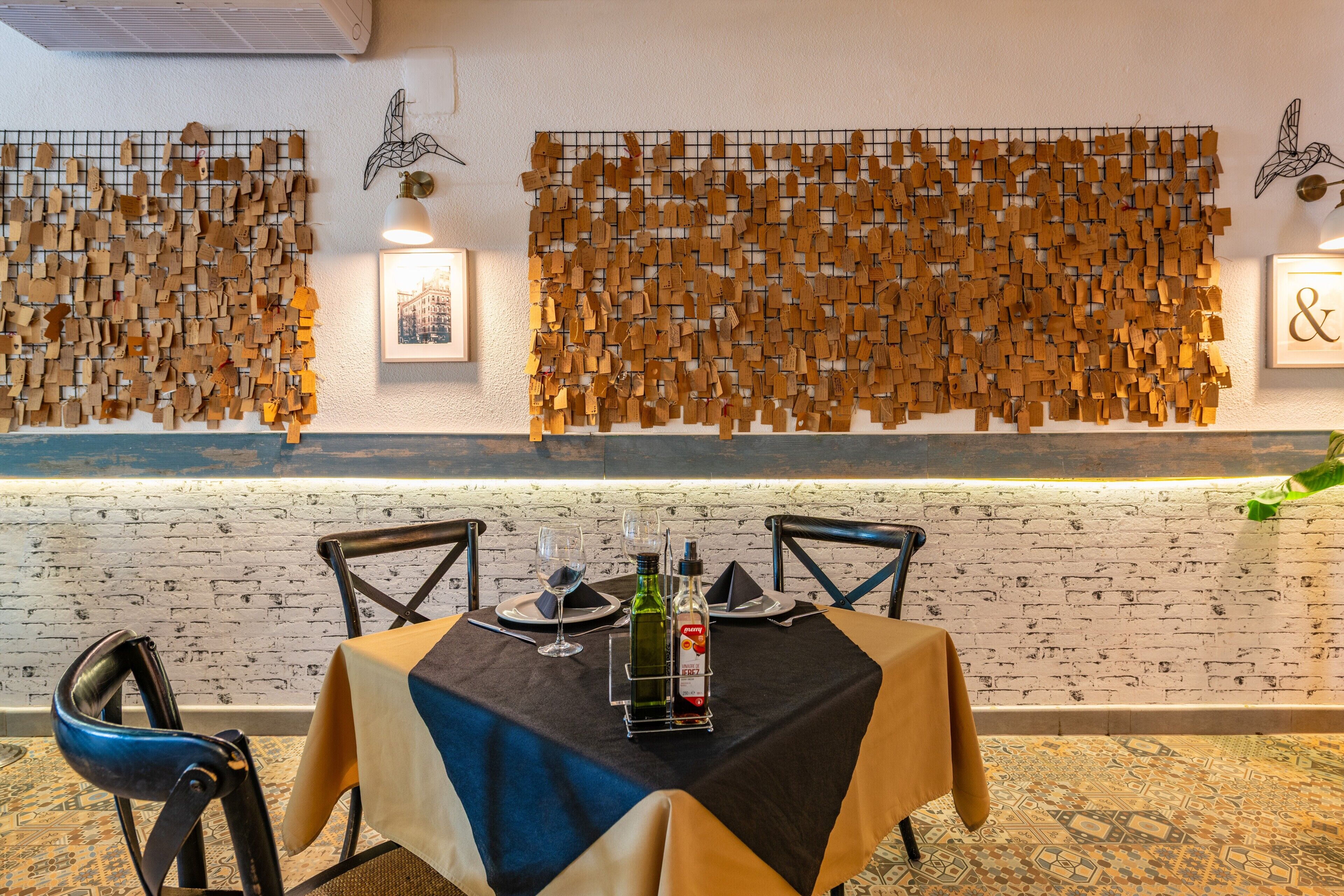 Un área de comedor con una mesa dispuesta para dos personas, una pared con un enrejado de etiquetas de madera y una pared de ladrillo con fotografías enmarcadas.