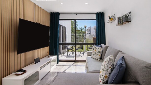 A modern living room with a flat-screen TV, a sofa, a coffee table, and a balcony with outdoor furniture.