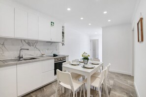 Una cocina moderna con armarios blancos, encimera de mármol y una zona de comedor con mesa y sillas blancas.