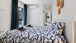 A bedroom with a patterned bedspread, a view of the kitchen, and a door leading to a balcony.