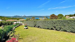 Un jardín ajardinado con seto, un banco de piedra y vista a un cuerpo de agua y colinas a lo lejos.