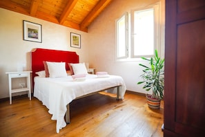 Une chambre avec un plafond en bois, un lit avec une literie blanche, une table de chevet, une commode, une fenêtre et une plante en pot.