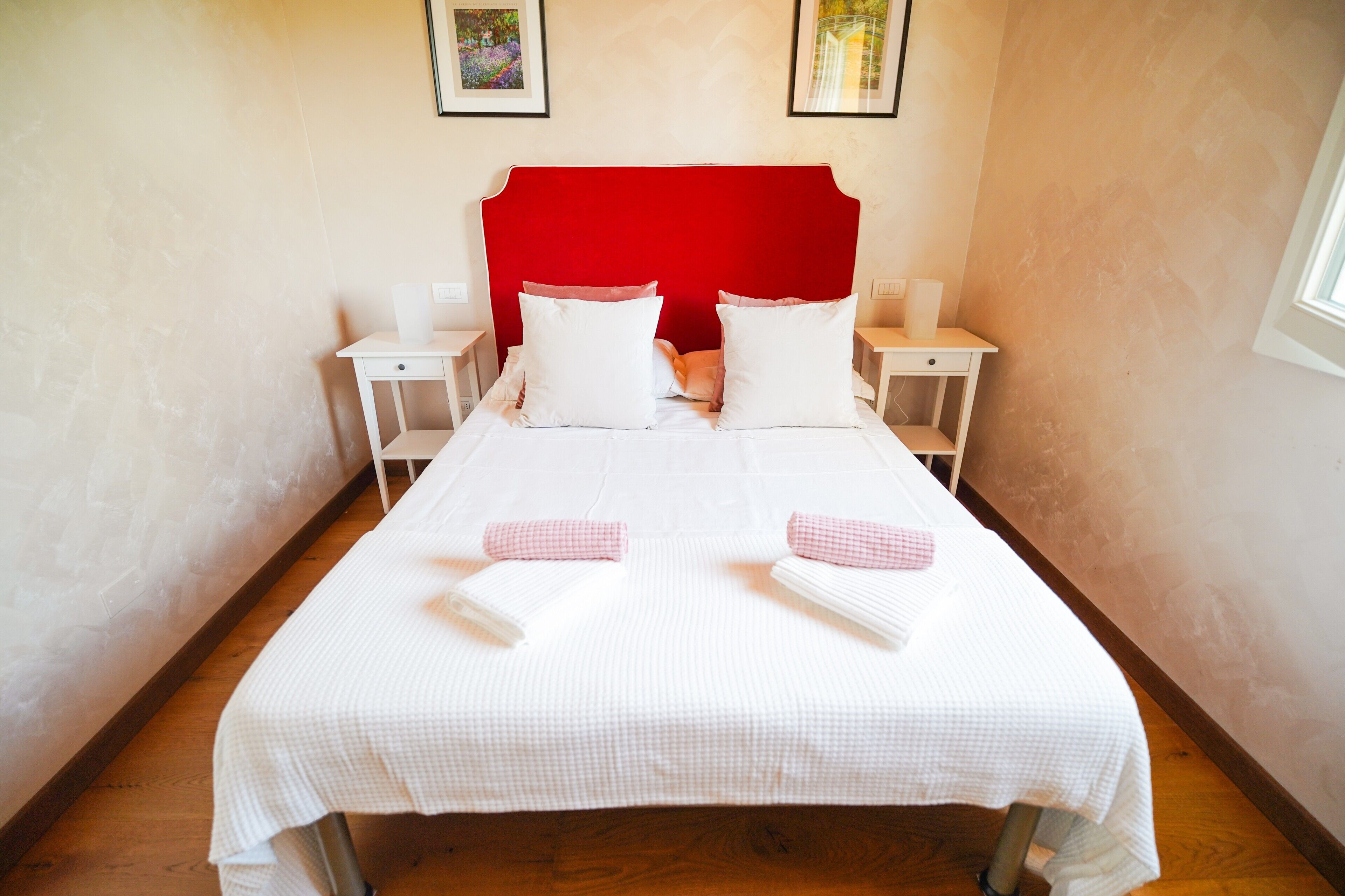 A neatly made bed with white linens and pink checkered towels, a red headboard, and bedside tables with lamps.
