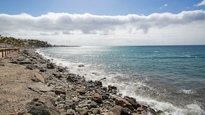 A rocky beach with a pier extending into the sea, clear skies, and distant buildings.