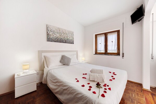 A neatly made bed with rose petals and a single rose, a bedside lamp, a small bedside table, and a painting on the wall.