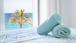 A bed with a patterned bedspread and a rolled towel on it, with a view of a palm tree and ocean through a window. - Apartment 'Sa Maniga' with Sea View, Wi-Fi and Air Conditioning (Sant Llorenç des Cardassar)
