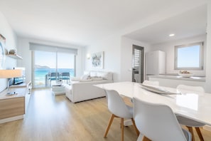 A modern kitchen with white appliances, a dining area with a white table and chairs, and a view of the sea through large windows.