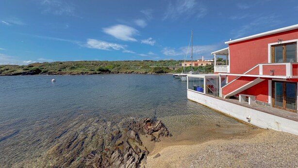 A coastal building with a red facade, a clear view of the water, and a rocky shoreline.