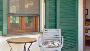 A window with green shutters, a white chair with a cushion, and a small round table.