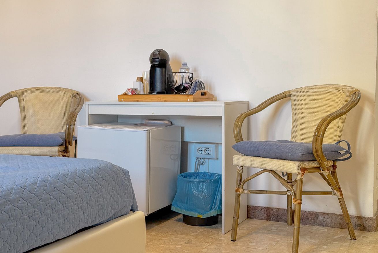 Una habitación pequeña con dos camas individuales, un escritorio con cafetera, un cubo de basura y dos sillones de mimbre.