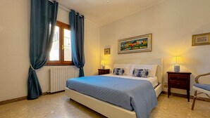 A bedroom with a bed, two bedside tables, a chair, a window with curtains, a radiator, and a painting on the wall.