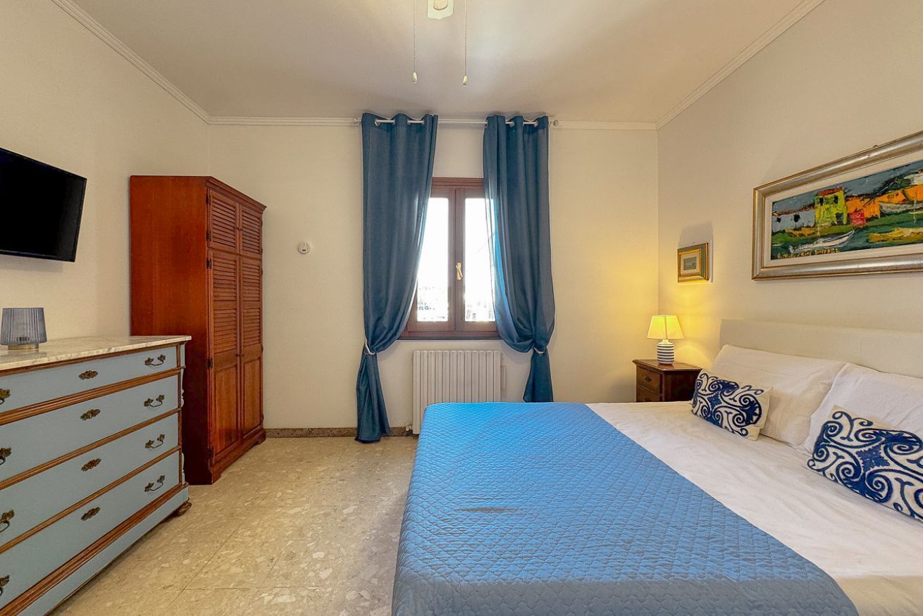 Ein Schlafzimmer mit einem Bett, einem Schminktisch, einem Fernseher, einem Fenster mit Vorhängen und einem Bild an der Wand.