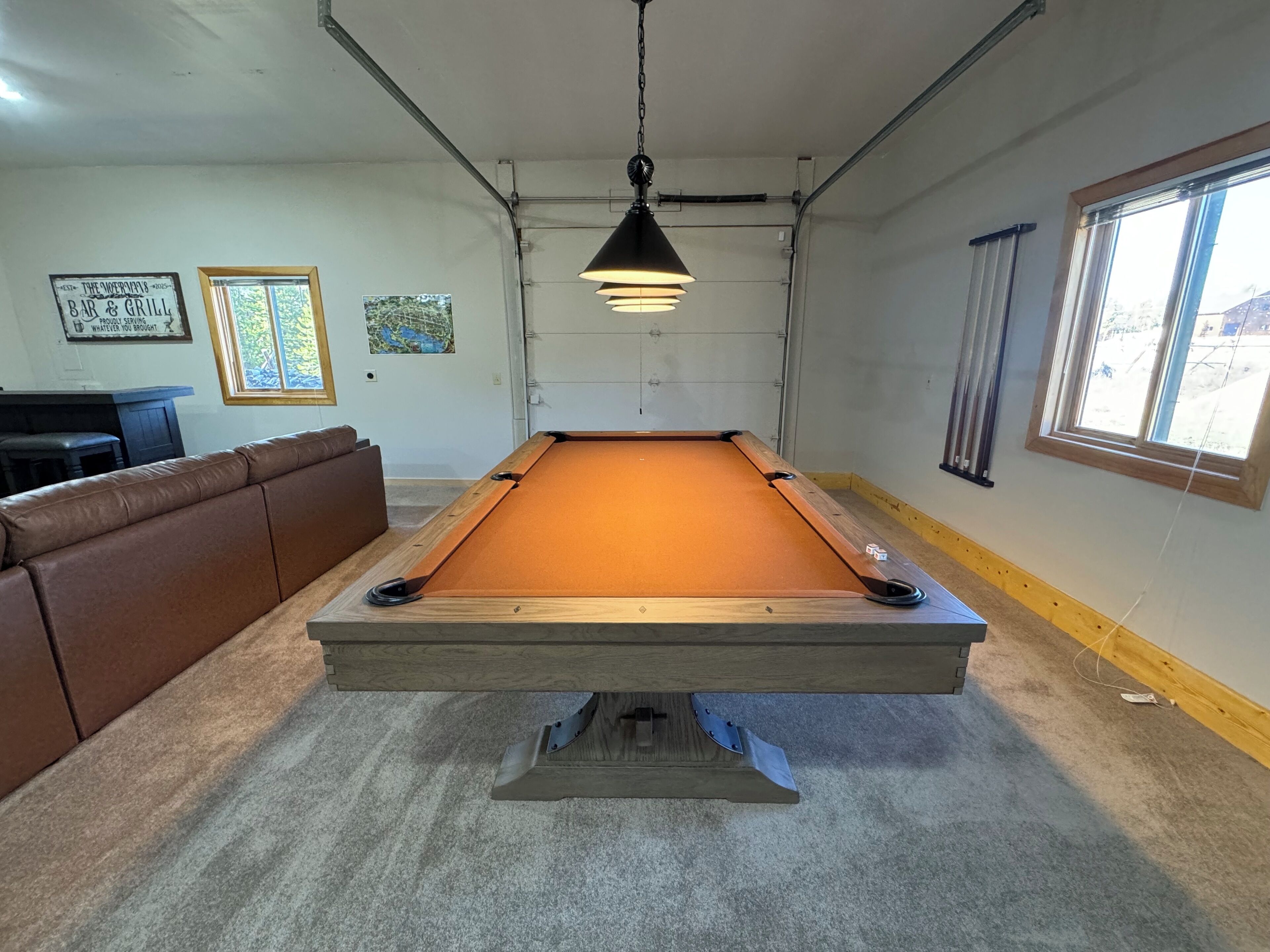 Een pooltafel in een kamer met een bruine leren bank, een hanglamp en een raam met jaloezieën.