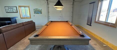 Een pooltafel in een kamer met een bruine leren bank, een hanglamp en een raam met jaloezieën.