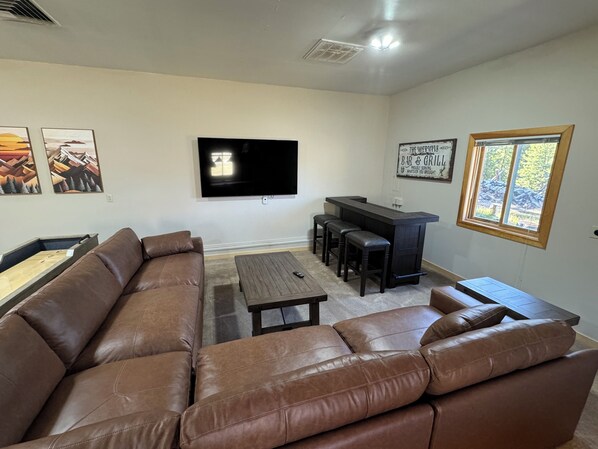 A living room with a brown leather sofa, a wooden coffee table, a TV mounted on the wall, and a bar area with stools.
