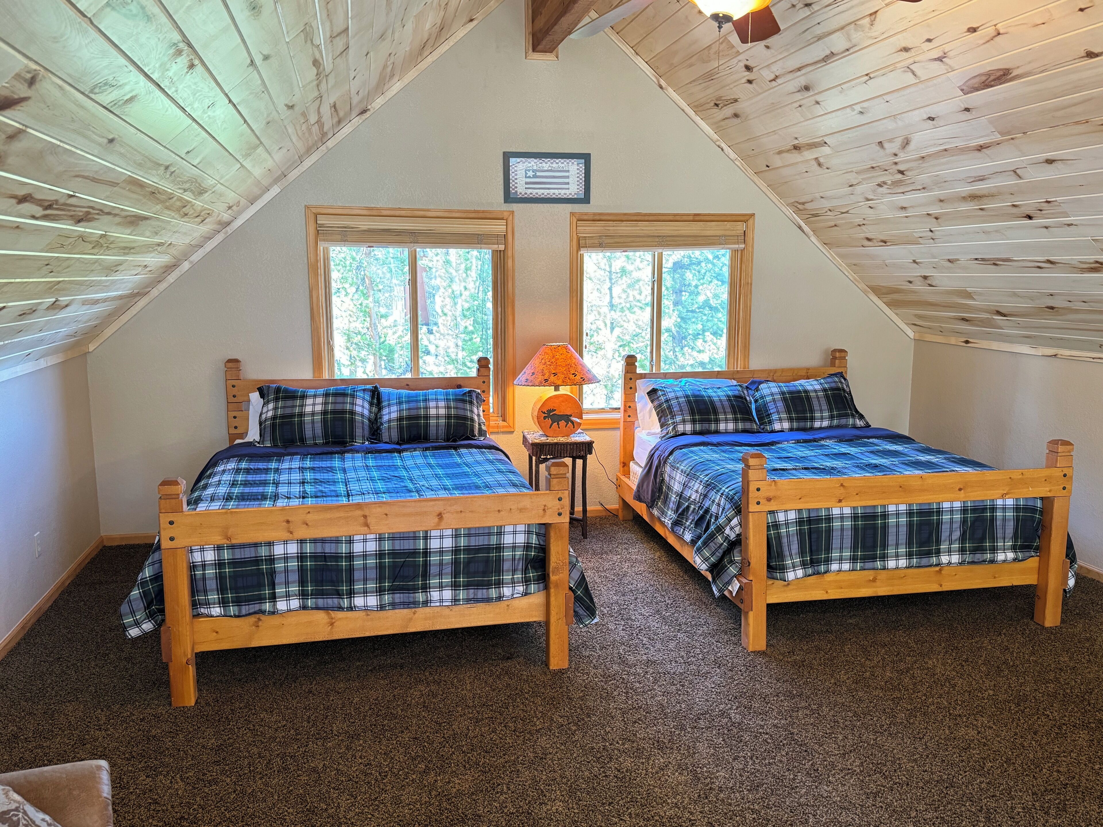 Ein gemütliches Schlafzimmer im Dachgeschoss mit zwei Holzbettchen, einem kleinen Tisch mit Lampe und großen Fenstern mit Blick auf Bäume.