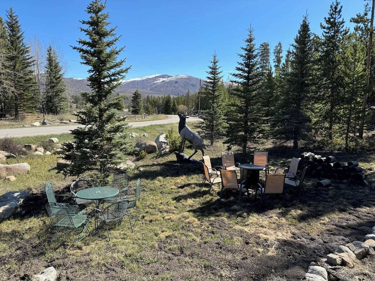 Ein rustikaler Sitzbereich im Freien mit Metallstühlen und einem Tisch, umgeben von Bäumen und einem Berg im Hintergrund.