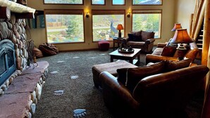 A cozy living room with a stone fireplace, leather furniture, and large windows offering a view of trees and a deer statue. - PICKLEBALL Paradise! HotTub/Huge GameRoom/Mtn views/Fido Friendly/best location! (Grand Lake)