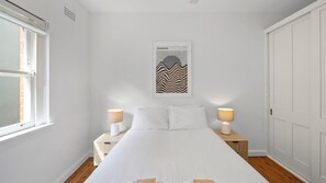 Ein Schlafzimmer mit einem Bett, zwei Nachttischen mit Lampen, einem Fenster und einer Tür.