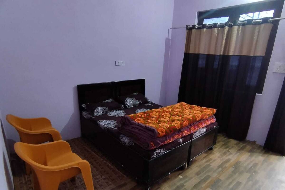 A bedroom with a bed, two orange chairs, a wooden floor, and a window with curtains.
