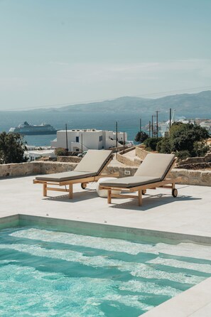 A pool area with two lounge chairs and a view of the sea and distant buildings. - Athera  Suites  Mykonos (Mykonos)