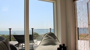 A room with a large sliding glass door leading to a balcony overlooking a body of water. There is a cushioned chair and a small table with binoculars on it. - "Valia" - 20m from the sea by Interhome (Sønder Stenderup)