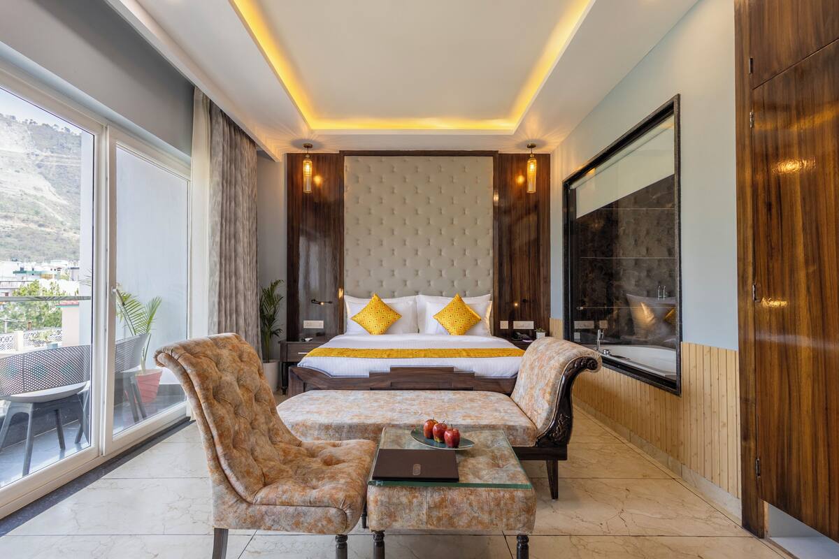 A bedroom with a large bed, a sofa, a coffee table, and a TV. There is a window with a view of the outside.