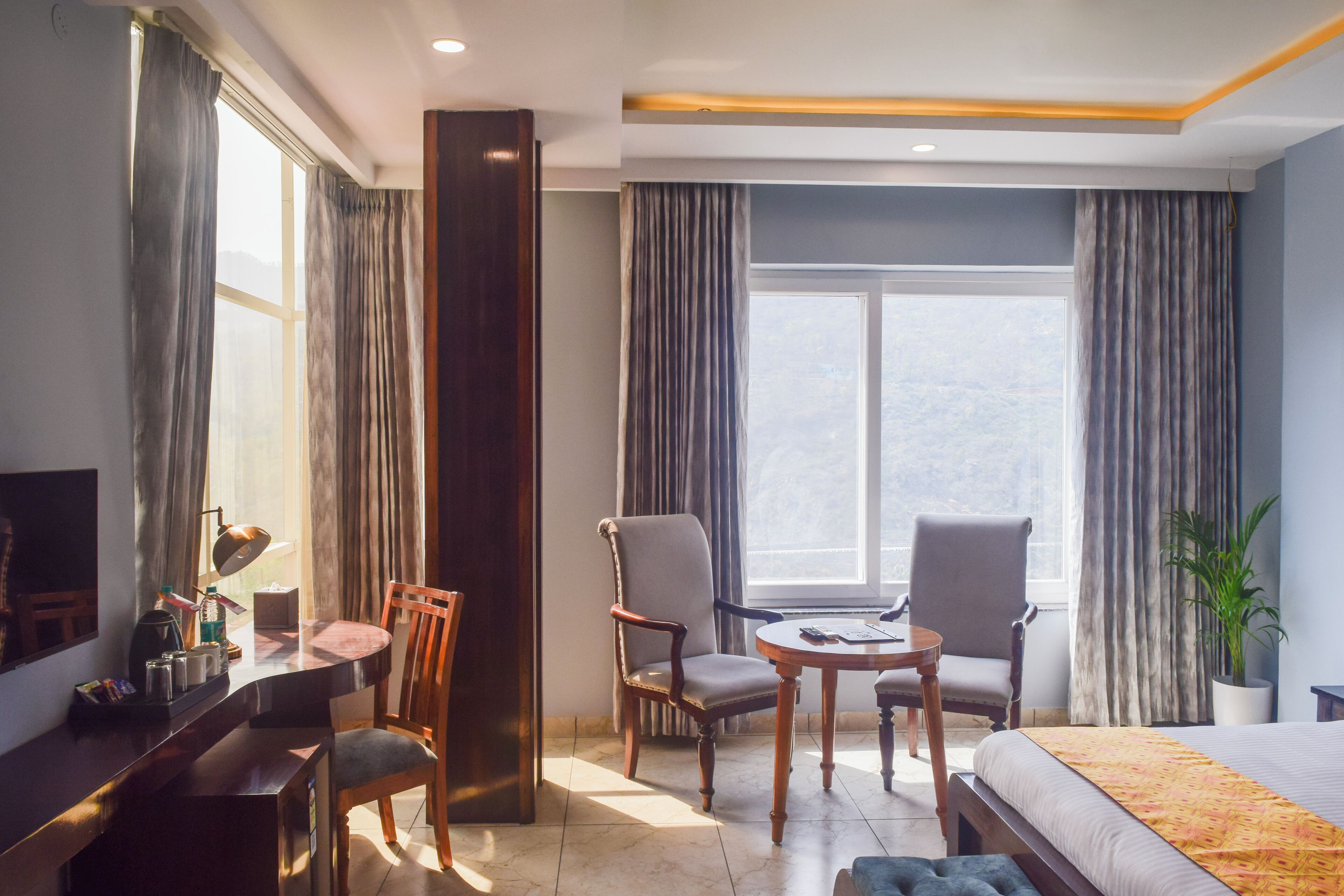 Habitació d'hotel amb llit, dues cadires, escriptori, làmpada i una finestra gran amb cortines.
