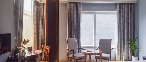A hotel room with a bed, two chairs, a desk, a lamp, and a large window with curtains.