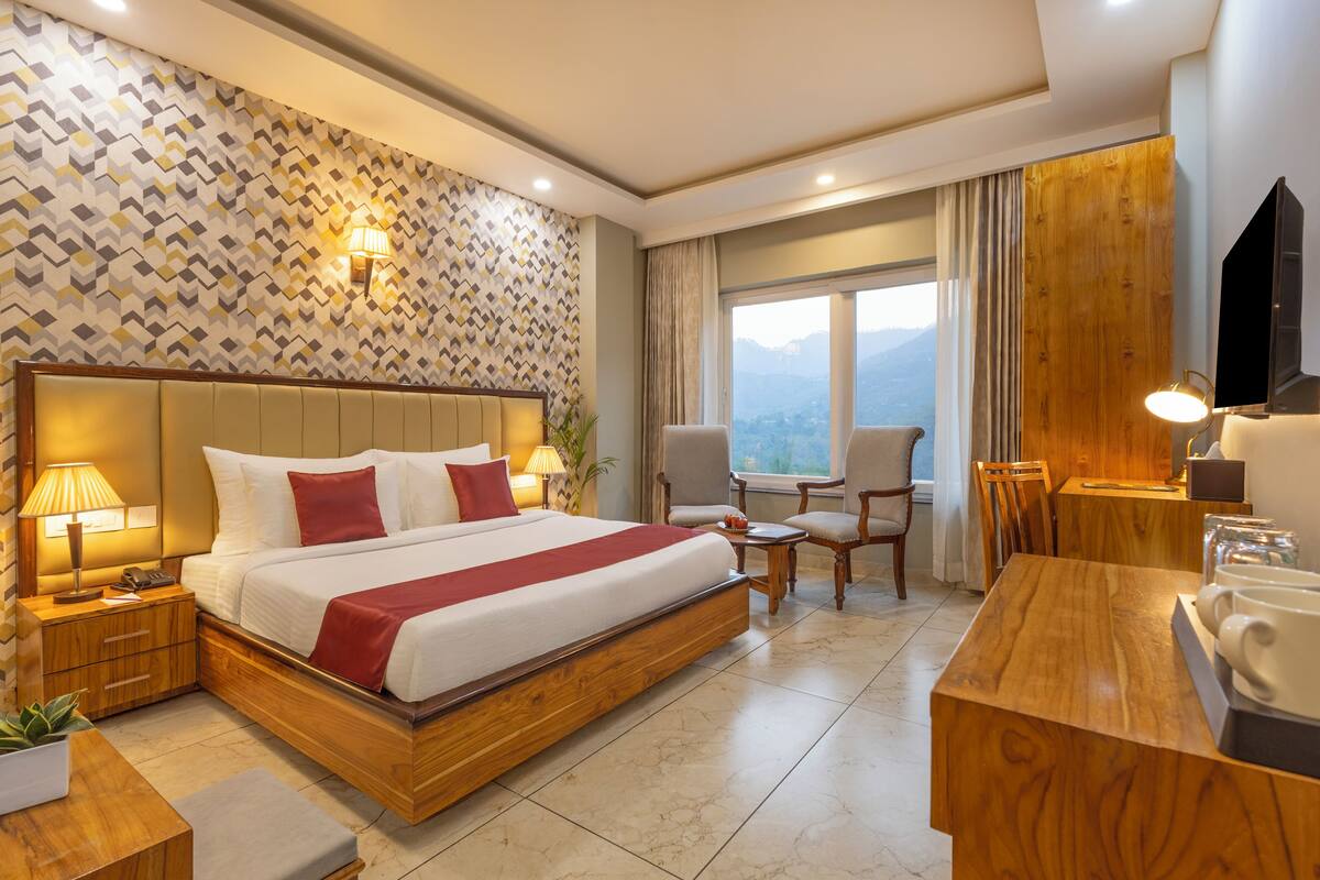 A hotel room with a large bed, a wooden headboard, a desk with a chair, a TV, and a window with a view of the mountains.