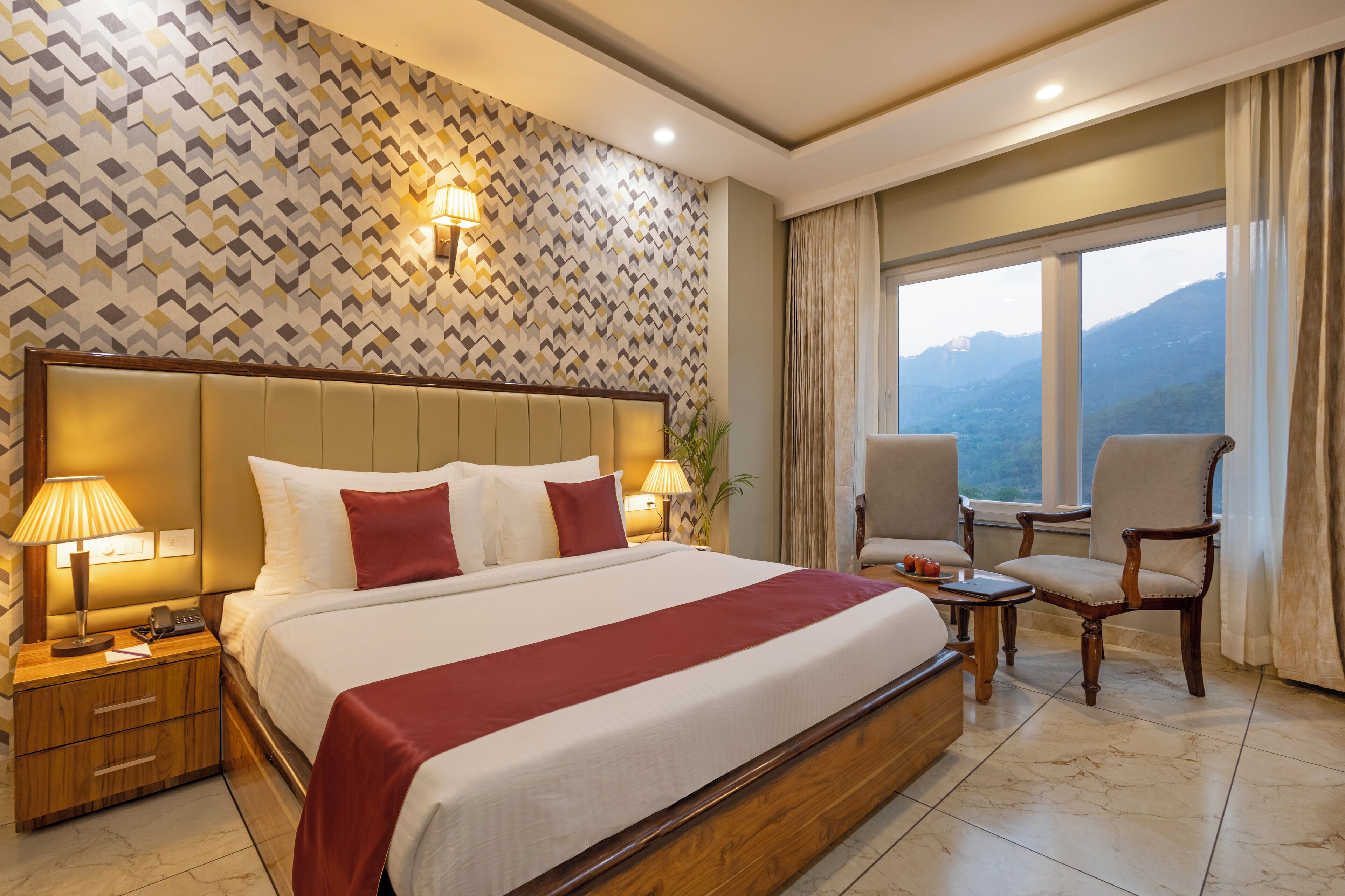 A hotel room with a large bed, two bedside lamps, a nightstand, a chair, a small table, and a window with a mountain view.