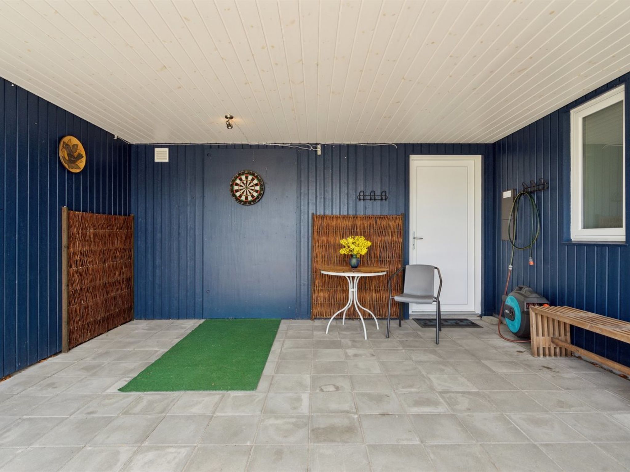 A room with a dartboard, a small table with a vase of flowers, a chair, and a wooden bench.