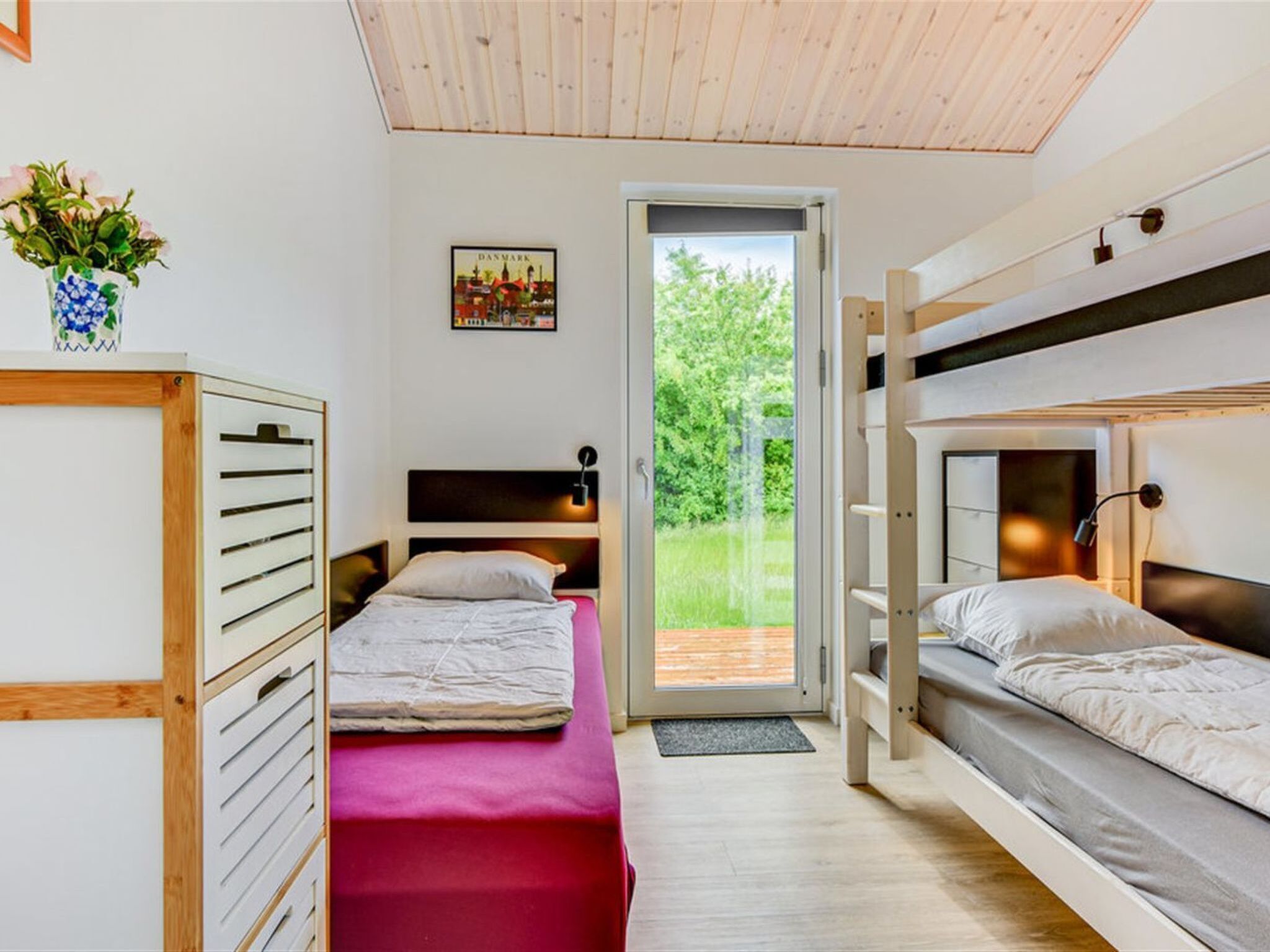 A room with two bunk beds, a wooden ceiling, a door leading to an outdoor area, and a small table with a vase of flowers.
