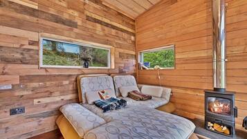 Relax in this serene retreat with a stove, two windows offering views of lush greenery, and a cozy bed with a patterned duvet.