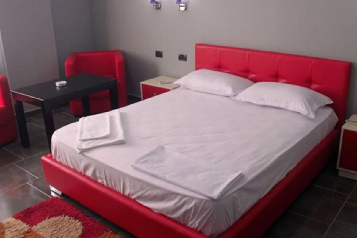 A hotel room with a red bed, white linens, a small table, red chairs, and a nightstand with a phone.